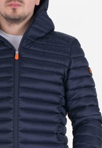 Marine gewatteerde jas met een rits aan de voorkant, kap en een oranje ritsen-trekker. Beschikt over een horizontaal gestikt ontwerp en een logo patch op de mouw.