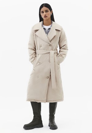Frau trägt einen beigen Trenchcoat mit Gürtel über geschichteten Oberteilen, kombiniert mit schwarzen kniehohen, klobigen Stiefeln, steht mit den Händen in den Taschen.