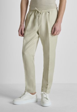 Osoba u bež hlačama s vezicama i bijelim tenisicama na vezanje, stoji na jednostavnoj svijetloj pozadini.