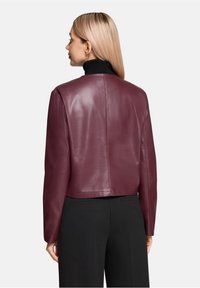 Kobieta o prostych blond włosach nosi krótka bordową skórzaną kurtkę na czarnym golfie i czarnych spodniach, stoi tyłem.