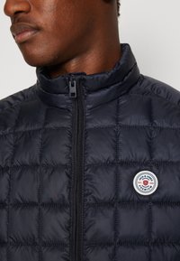 Marinblå quiltad pufferjacka med hög krage, med dragkedjestängning och cirkulär logotyp på vänster bröst. Slät, glänsande yta.