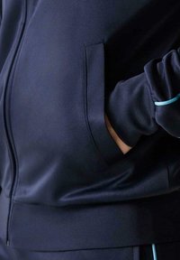 Chaqueta deportiva con cremallera en color azul marino, hecha de tela suave, con un bolsillo lateral y detalles en azul claro en los puños.