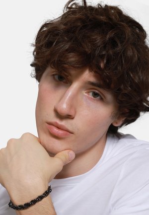 Curly brown hair, pale complexion, wearing a white T-shirt. Black chain bracelet on wrist; hand resting on chin, neutral expression.