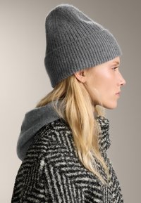 Blondynka w profilu nosząca szary, dziany beret oraz fakturowany czarno-biały płaszcz z szarym kapturem.