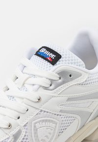 Weiße sportliche Sneaker aus Mesh- und Leder-Materialien, mit einem auffälligen schwarzen Etikett mit der Aufschrift 'Blauer', silbernen Akzenten und weißen Schnürsenkeln.