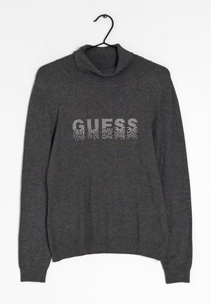 Pull en maille gris avec un col roulé, arborant le mot "GUESS" en strass sur le devant. Manches et ourlet extensibles. Suspendu à un crochet noir.