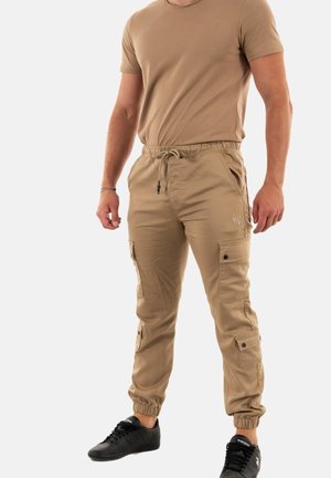 Pantaloni cargo beige con polsini elasticizzati, cintura regolabile, numerose tasche e dettagli cuciti, abbinati a una T-shirt color sabbia e sneaker nere.