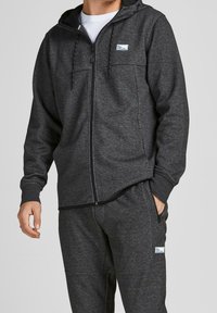 Sudadera con cremallera en gris oscuro, fabricada en un tejido suave, con un bolsillo tipo canguro, capucha con cordón y parche de logo en el pecho. Combinada con unos joggers a juego.