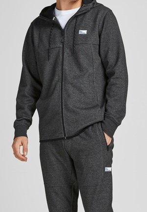 Hoodie zippé gris foncé en tissu doux avec une poche kangourou, un capuchon à cordon et un patch logo sur la poitrine. Associé à un pantalon de jogging assorti.