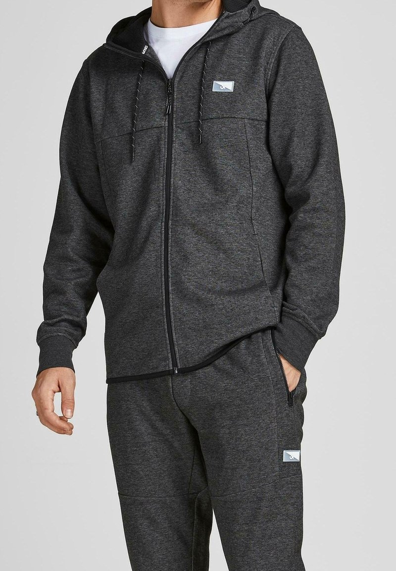 Sudadera con cremallera en gris oscuro, fabricada en un tejido suave, con un bolsillo tipo canguro, capucha con cordón y parche de logo en el pecho. Combinada con unos joggers a juego.