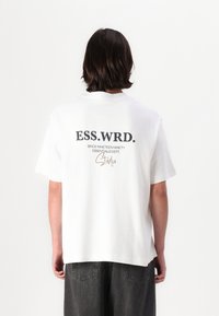 Bijela pamučna majica s okruglim izrezom, na leđima s otisnutim tekstom u crnoj i smeđoj boji: "ESS.WRD. OD DEVETNAESTDEVEDESET STUDIO."