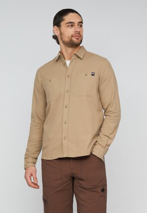 Jonge man met donker haar naar achteren gebonden, die een beige overhemd met knopen en een bruine broek draagt, kijkt opzij met één hand in de zak.