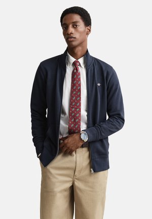 Veste zippée marine en matériau doux, associée à une chemise blanche et une cravate rouge à motifs, ainsi qu'un pantalon kaki et une montre argentée.