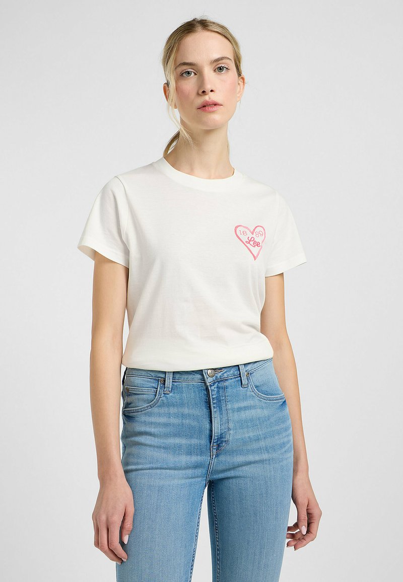 Hvid bomulds t-shirt med korte ærmer, prydet med et pink hjerte grafik og "Lee" tekst. Kombineret med lyseblå denim jeans.