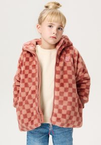 Roze geruite fleece jas met capuchon, voorzien van een roze rits en een badge. Gedragen over een crèmekleurig topje en blauwe jeans. Zachte textuur.