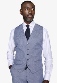 Hombre con barba gris que lleva un chaleco de cuadros azul claro y pantalones, camisa blanca y corbata negra, mirando de lado con las manos en los bolsillos.