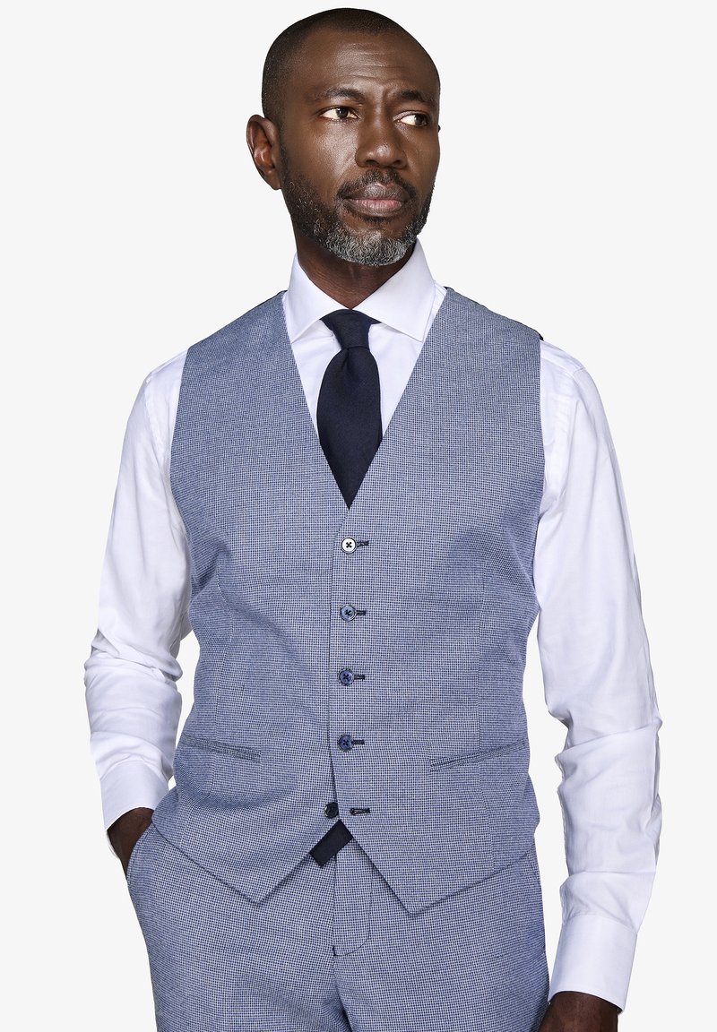 Hombre con barba gris que lleva un chaleco de cuadros azul claro y pantalones, camisa blanca y corbata negra, mirando de lado con las manos en los bolsillos.