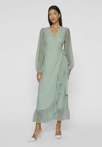 Myntgrønn innpakningskjole med prikkete mønster, lange gjennomsiktige ermer, volangkant og knytelukking i midjen. Strukturerte stoffoverflate.