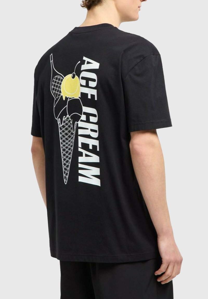 En person som bär en svart tröja med en stor glasskon-överskrift och texten "ACE CREAM" på baksidan, mot en enkel grå bakgrund.