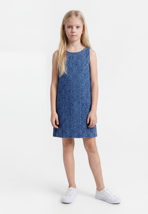 Niña joven con pelo largo y rubio, vestido azul sin mangas con estampado y zapatillas blancas, de pie frente a un fondo blanco liso.