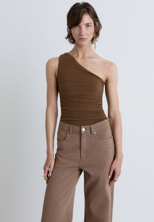 Woman with short brown hair wearing a brown one-shoulder top and high-waisted brown pants standing against a plain light background.