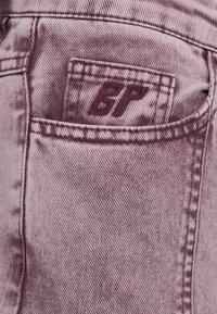 Gros plan sur la poche arrière d'un jean en denim violet avec une petite poche à l'intérieur, ornée des initiales brodées "BF" en violet foncé.