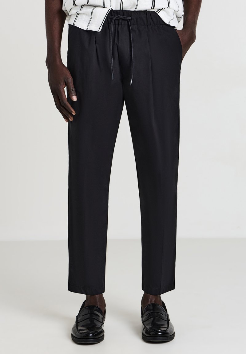 Uomo con pantaloni neri a coulisse e mocassini neri lucidi, in piedi con le mani nelle tasche contro uno sfondo semplice.