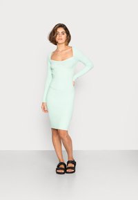 Vestido verde menta com textura em relevo, mangas longas e decote quadrado, de corte ajustado, combinado com sandálias pretas com uma faixa larga.