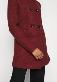 Abrigo de mezcla de lana rojo con un acabado texturizado, parte frontal de doble botonadura, botones negros y bolsillos laterales, con una silueta ligeramente en A.