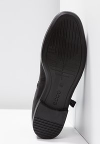 Zapato de cuero negro con suela de goma texturizada, que presenta un perfil bajo y diseño minimalista. Logo grabado en el talón.