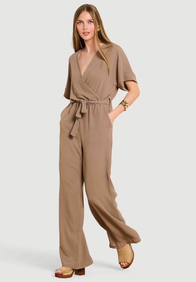 Giovane donna che indossa una tuta larga color sabbia con top incrociato e cintura, posando con una mano in tasca e sandali beige con tacco.
