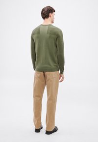 Zöld kötött pulóver hosszú ujjakkal, bordázott mintával a vállakon, bézs nadrággal és fekete cipővel párosítva, hátulnézetből.