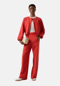 Rote Satin-Bomberjacke mit weißem Oberteil und roter Hose. Das Modell hält eine kleine beige Handtasche und trägt weiße Sneaker.