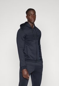 Marineblå hettejakke med glidelås, teksturert brystpanel, frontlommer og justerbar hette med snor. Laget av mykt, elastisk stoff.