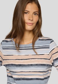Junge Frau mit langen braunen Haaren, die ein pastellgestreiftes Hemd mit horizontalen blauen, beigen, grauen und schwarzen Streifen auf einem hellgrauen Hintergrund trägt.
