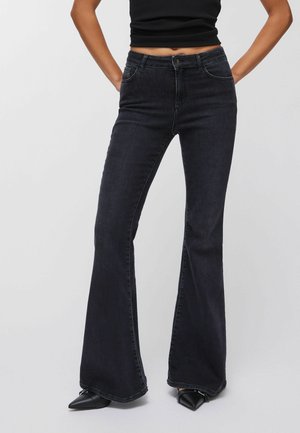 Žena v čiernych džínsach s vysokým pásom a rozšírenými nohavicami, čiernom bezrukávovom krátkom topu a čiernych špicatých topánkach, stojaca s rukami v bok.