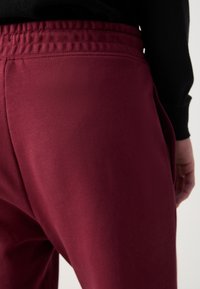 Burgunderfarbene Jogginghose mit elastischem Bund, seitlichen Taschen und glatter Textur. Der Stoff ist weich und dehnbar für maximalen Komfort.