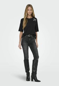 Tricou negru cu mânecă scurtă, având un detaliu decorativ sub formă de fundiță, asortat cu blugi închisi la culoare și cizme negre din piele până la genunchi, cu tocuri groase.