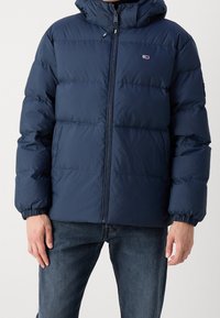 Marinblå puffjacka med huva, med horisontella kviltade sektioner, dragkedjestängning och elastiska cuffar. Materialet verkar slätt och isolerat.