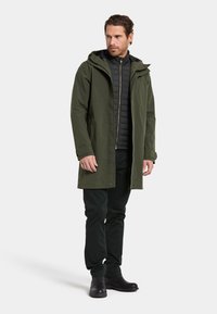 Oliven grøn vandtæt parka med lynlås, stor hætte og justerbare manchetter, over en sort quiltet inderforing, parret med mørke bukser og støvler.