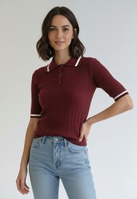 Bordowa dzianinowa koszulka polo z teksturowanym wzorem, bia�łe rękawy z paskami i listwa z trzema guzikami. W zestawie z jasnoniebieskimi dżinsami.