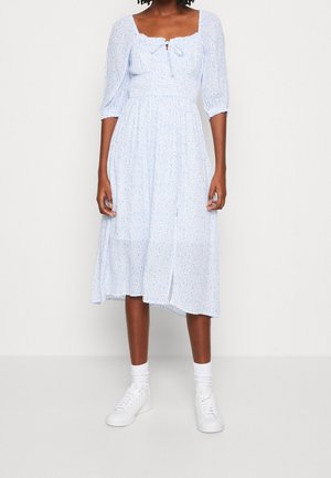 Vrouw draagt een lichtblauwe midi-jurk met bloemenprint en opgeblazen driekwartmouwen, gecombineerd met witte sokken en witte sneakers.