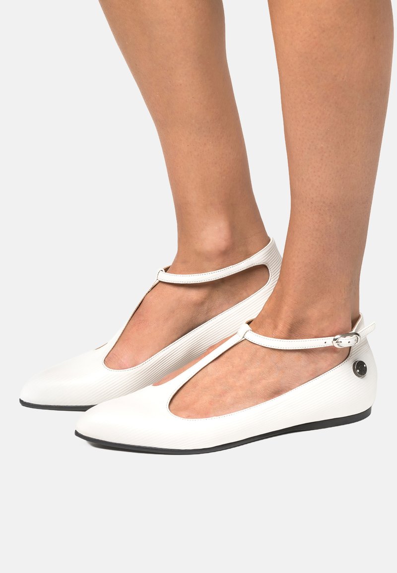 Scarpe bianche flat con una tomaia testurizzata, design a T e cinturino alla caviglia. Presentano una punta rotonda e sottili accenti neri sulla suola.