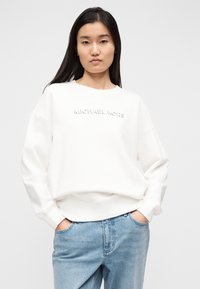 Άσπρο φούτερ με ριμπ μανσέτες και τελείωμα, με ανάγλυφο λογότυπο "MICHAEL KORS" στο στήθος. Συνδυάζεται με ανοιχτό μπλε τζιν παντελόνι.