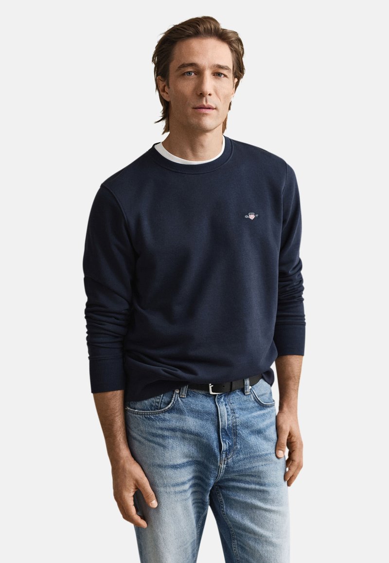 Marineblå sweatshirt med ribstrikkede ærmekanter og halsudskæring, prydet med en lille broderet logo. Båret med lyseblå jeans og et sort bælte.