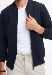 Granatowa kurtka bomber z teksturowanym, pikowanym wzorem, ściągaczami na mankietach i otwartym zamkiem błyskawicznym. Noszona na białej koszulce, w połączeniu z jasnobeżowymi spodniami.