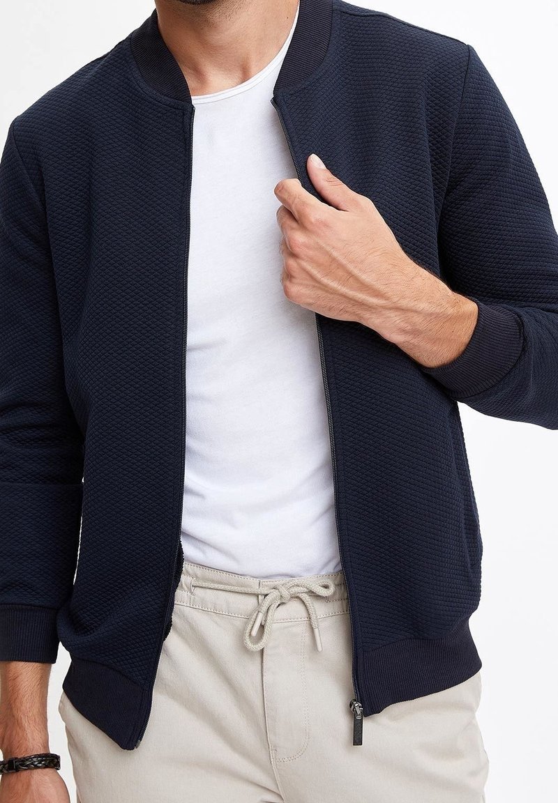 Granatowa kurtka bomber z teksturowanym, pikowanym wzorem, ściągaczami na mankietach i otwartym zamkiem błyskawicznym. Noszona na białej koszulce, w połączeniu z jasnobeżowymi spodniami.