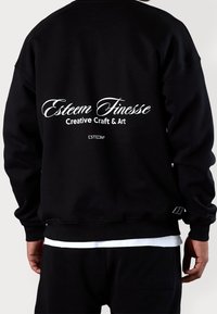Uomo che indossa una felpa nera con la scritta bianca "Esteem Finesse Creative Craft & Art" stampata sulla schiena, abbinata a pantaloni neri.