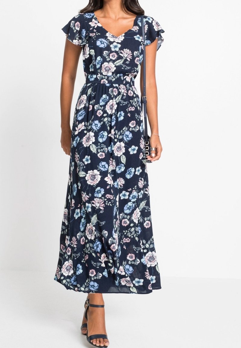 Femme portant une robe longue fleurie bleu marine avec des manches volantées, un col en V et des sandales à bride à la cheville, portant un petit sac bandoulière.