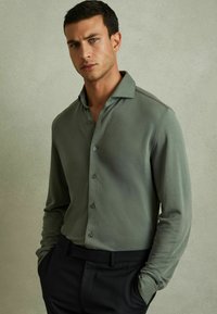 Cămașă lungă cu mâneci, de culoare verde măsliniu, cu guler și închidere cu buton, având o textură netedă și un design ajustat, asortată cu pantaloni negri.